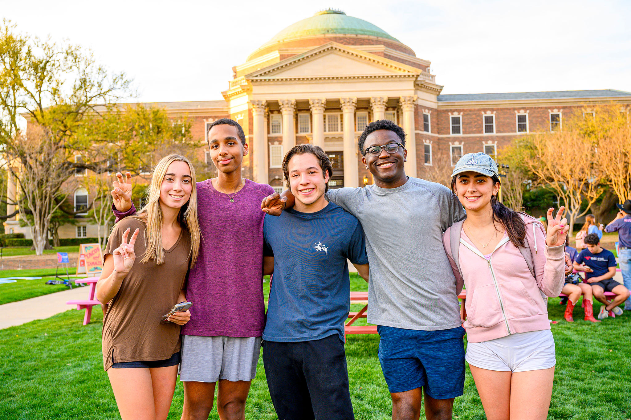 five students on lawn holding pony up hand signal in front of dallas hall at outdoor event