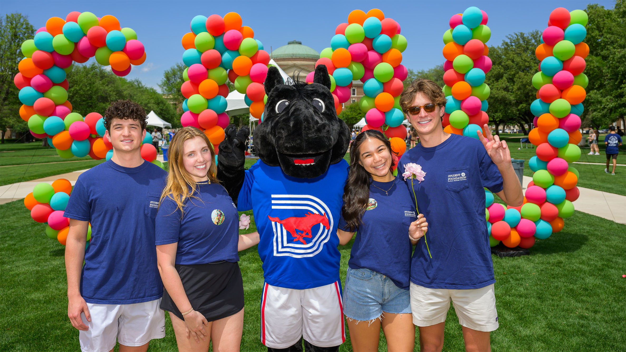 students standing with Peruna mascot outside in front of bright color balloon sculpture in letter S M U