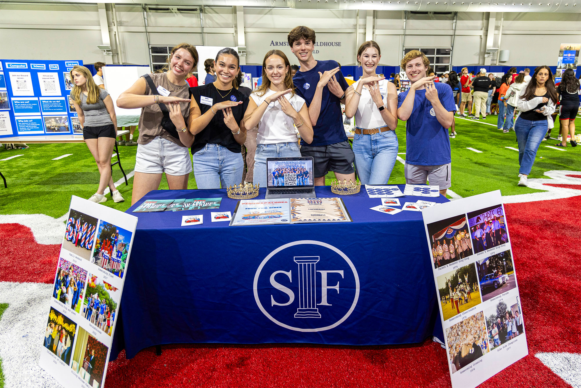 students standing behind table with blue tablecloth with paper and posters on table at a student club fair